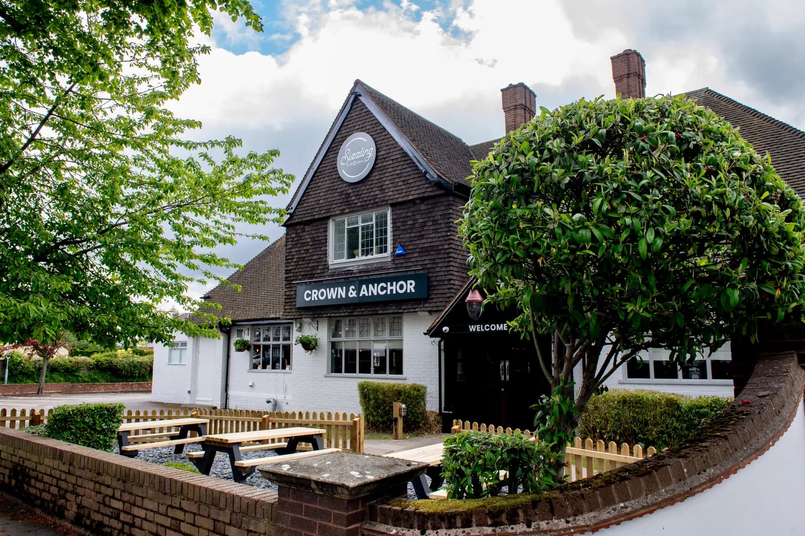 The white and dark brown exterior of the Crown & Anchor pub is shown with outdoor seating and a brick fence in the foreground.