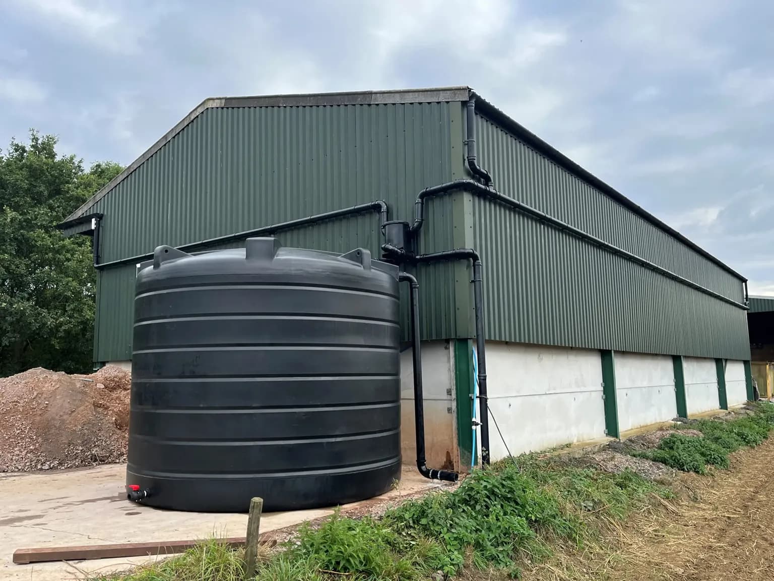 A large black water tank stands next to a green corrugated metal agricultural building with white concrete siding, under a cloudy sky.