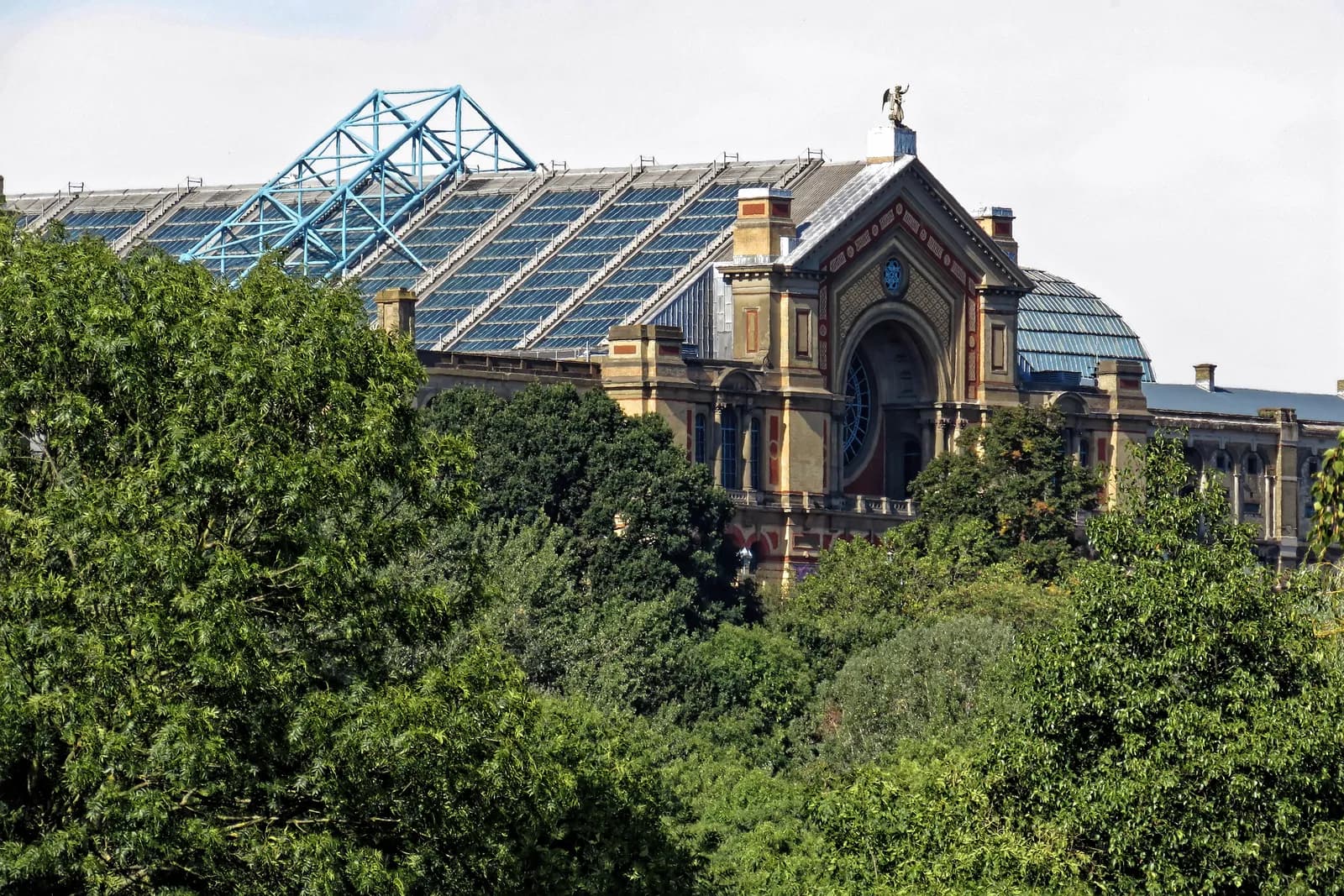 A grand, ornate building with a glass roof and blue structural elements rises above a dense canopy of lush green trees under a bright sky.