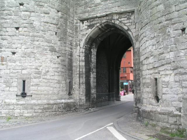 A large stone archway cuts through a massive stone wall, revealing a street and a red brick building on the other side.