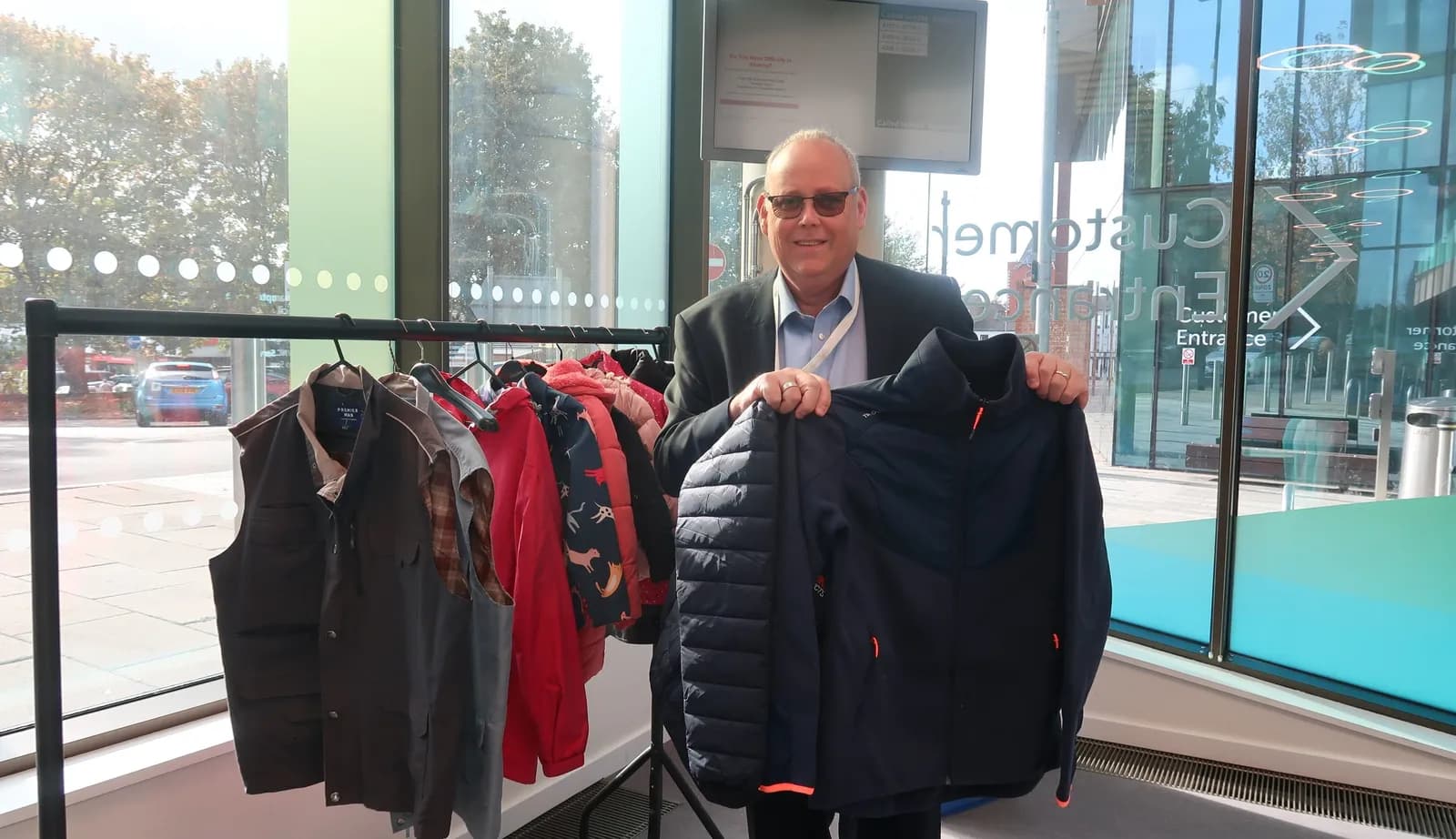 A man in a suit jacket and sunglasses stands next to a clothing rack, holding up a dark blue puffer jacket, with a glass building and trees in the background.