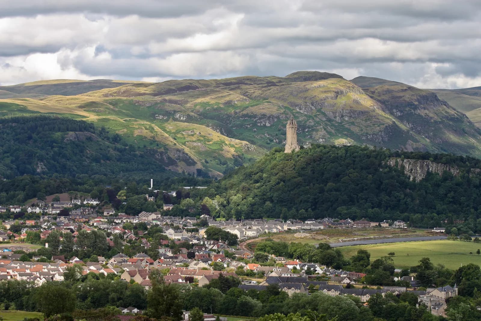 A towering stone monument is visible on a tree-covered hillside, with a town nestled in the valley below and rolling green hills under a cloudy sky.
