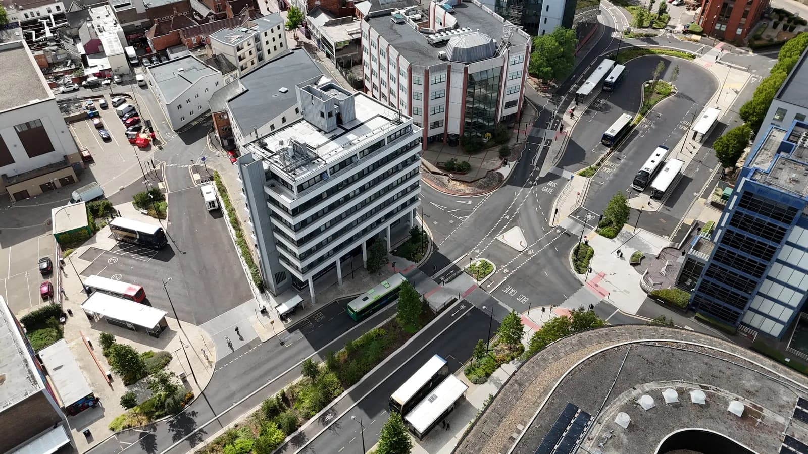 An aerial view of a busy urban area featuring multiple bus stops with several buses, surrounded by modern buildings and a complex network of roads with cars.