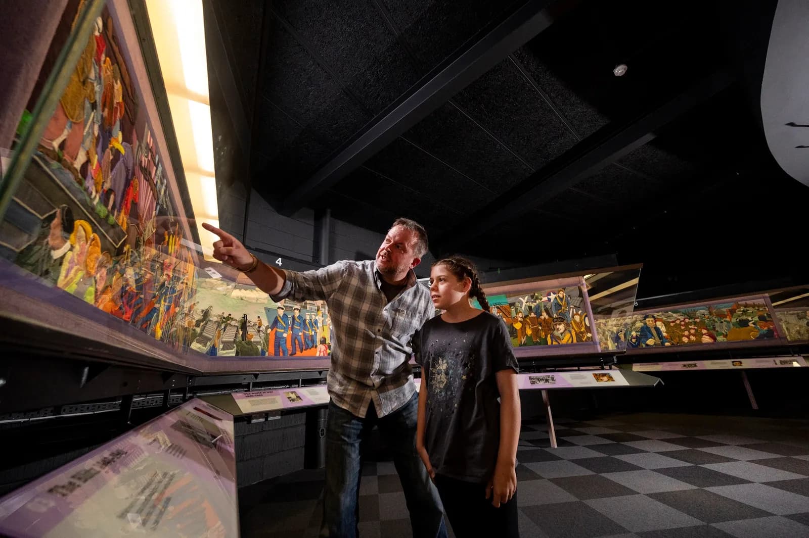 A man and a young girl standing in front of a curved mural display inside a dark museum, with the man pointing towards a section of the artwork.