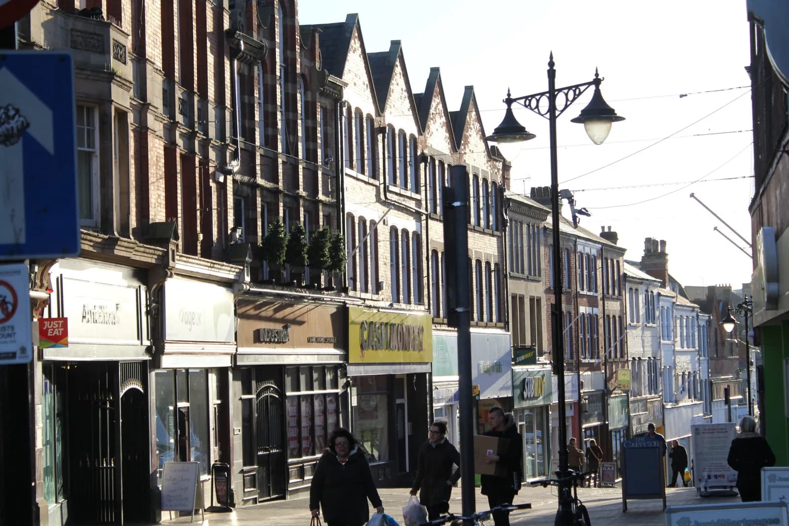 People walk past shops and residential buildings lining a street with a lamp post and street signs on a bright day.