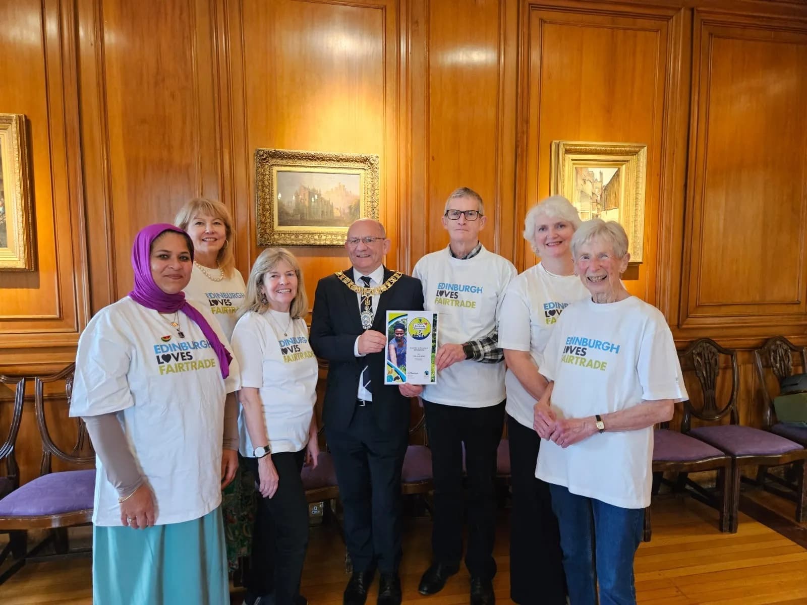 A group of people, some wearing "Edinburgh Loves Fairtrade" t-shirts, stand with a man in a ceremonial chain holding an award certificate.