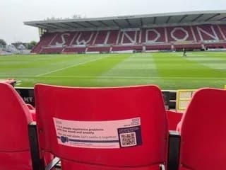 A close-up of an empty red stadium seat with a white sticker on its back, in front of a green soccer field and the stadium stands with "SWINDON" spelled out in large white letters.