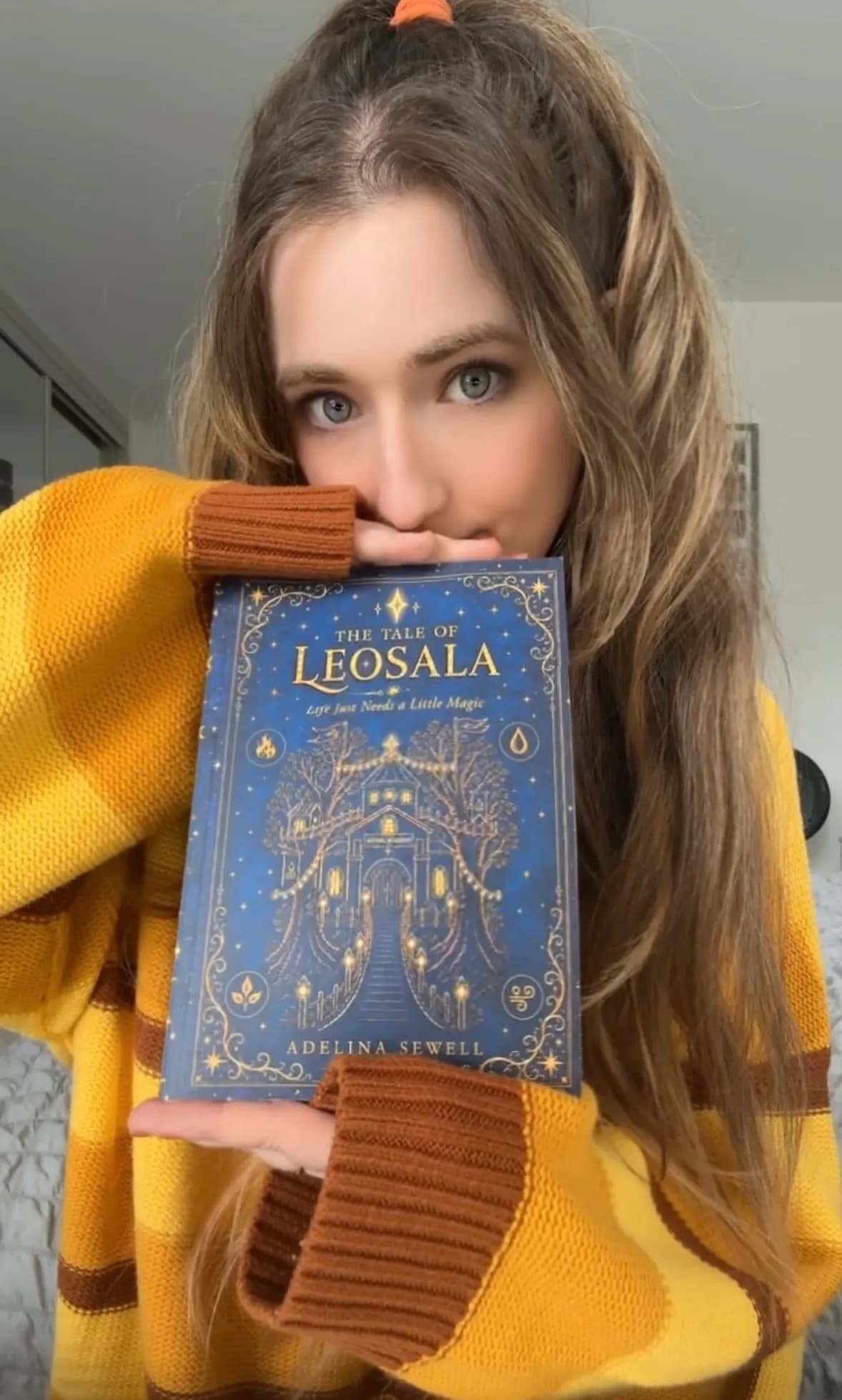A woman in a striped yellow and brown sweater holds a blue book titled "The Tale of Leosala" up to her face, partially obscuring her nose and mouth.