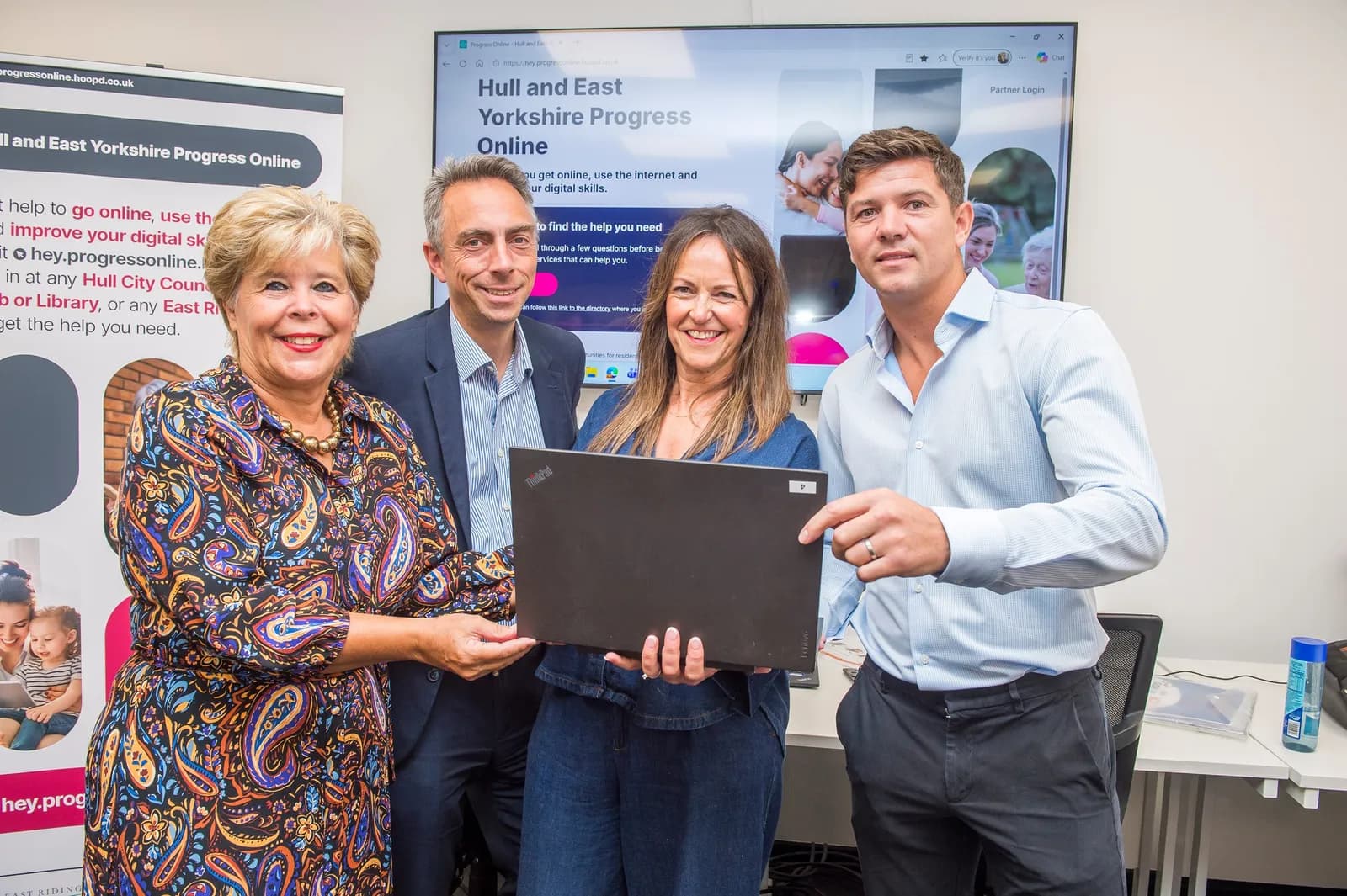 Four adults, two men and two women, stand together in front of a digital screen and a banner, holding a laptop computer.