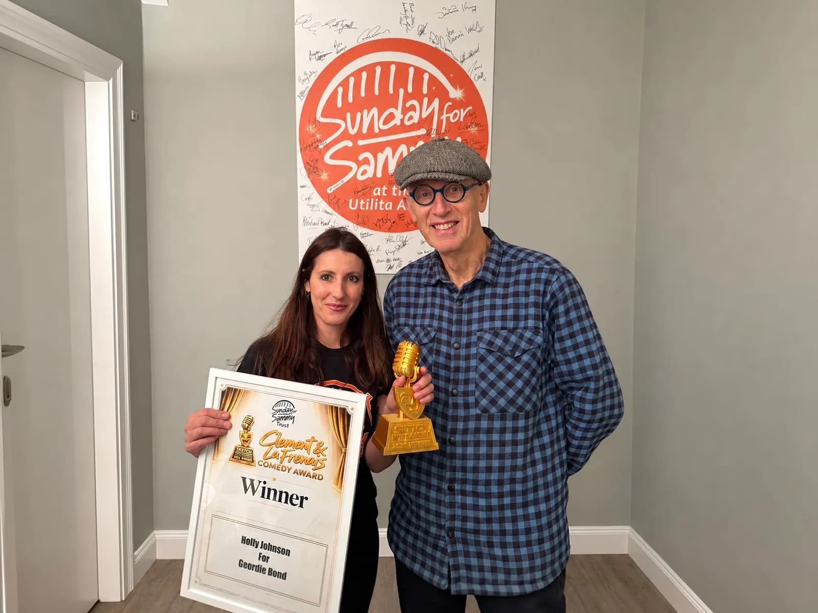 A woman and a man smile at the camera, holding a framed award certificate and a golden microphone trophy.