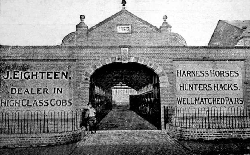 A black and white photo of a brick building with an arched entrance leading to a long, covered walkway with stalls, and a young boy standing near the entrance.