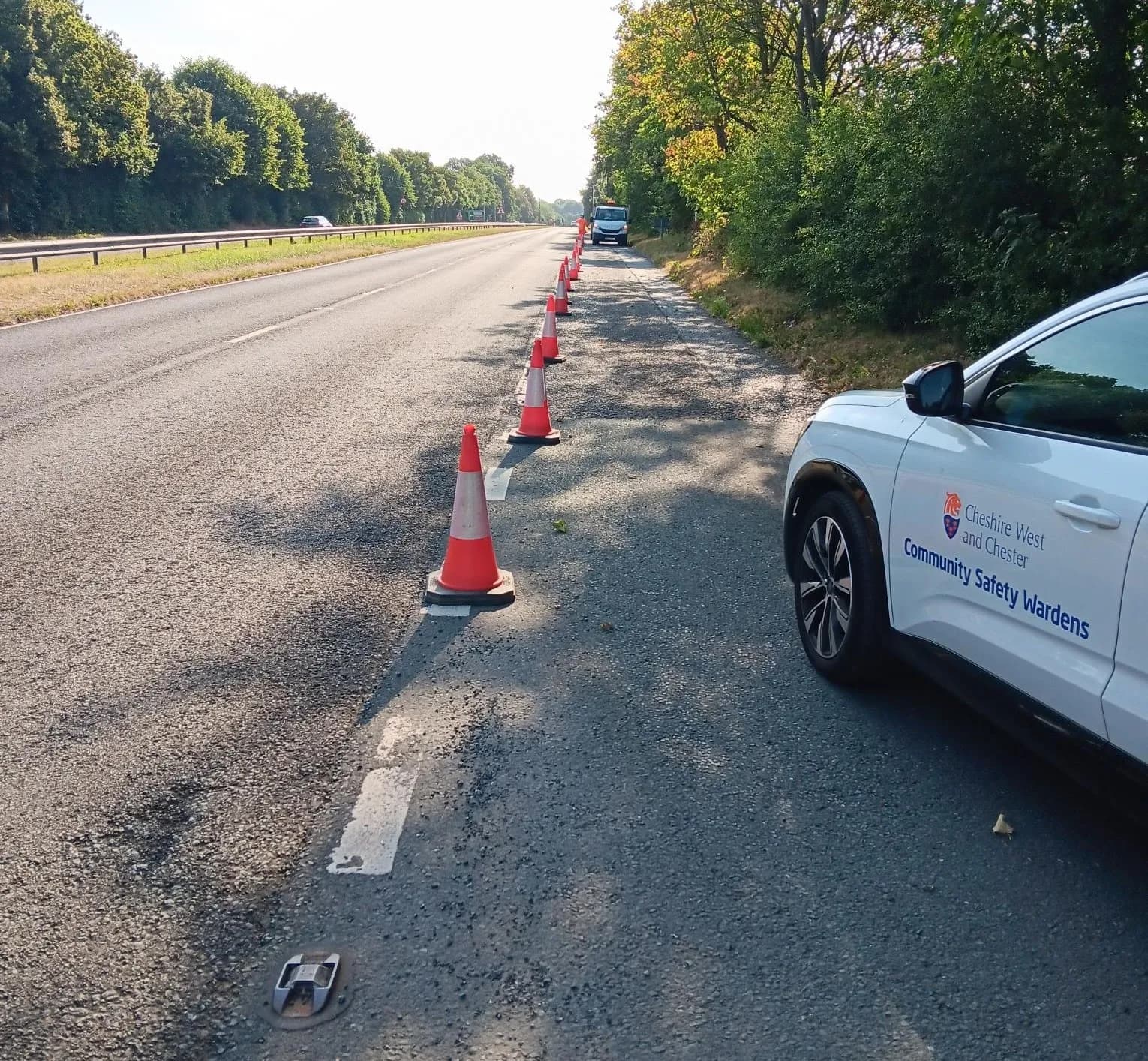 A white 'Community Safety Wardens' vehicle is parked on the shoulder of a tree-lined highway, next to a row of orange traffic cones.