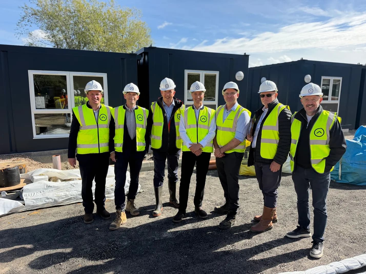 A group of six men in hard hats and high-visibility vests stand outside dark grey modular buildings under a bright sky.