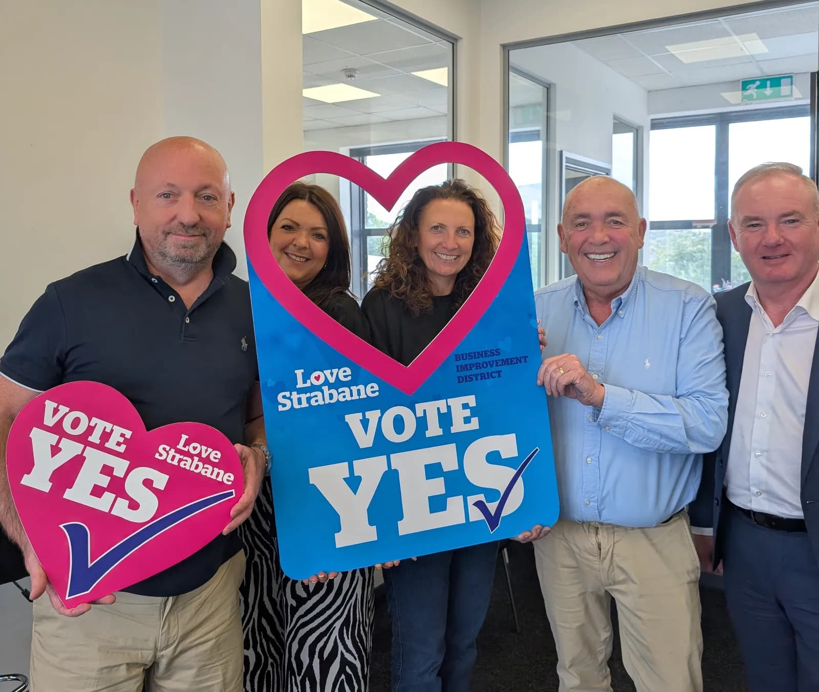 Five people, three men and two women, smiling and holding up two heart-shaped signs, one pink and one blue, that say "Vote Yes" and "Love Strabane."