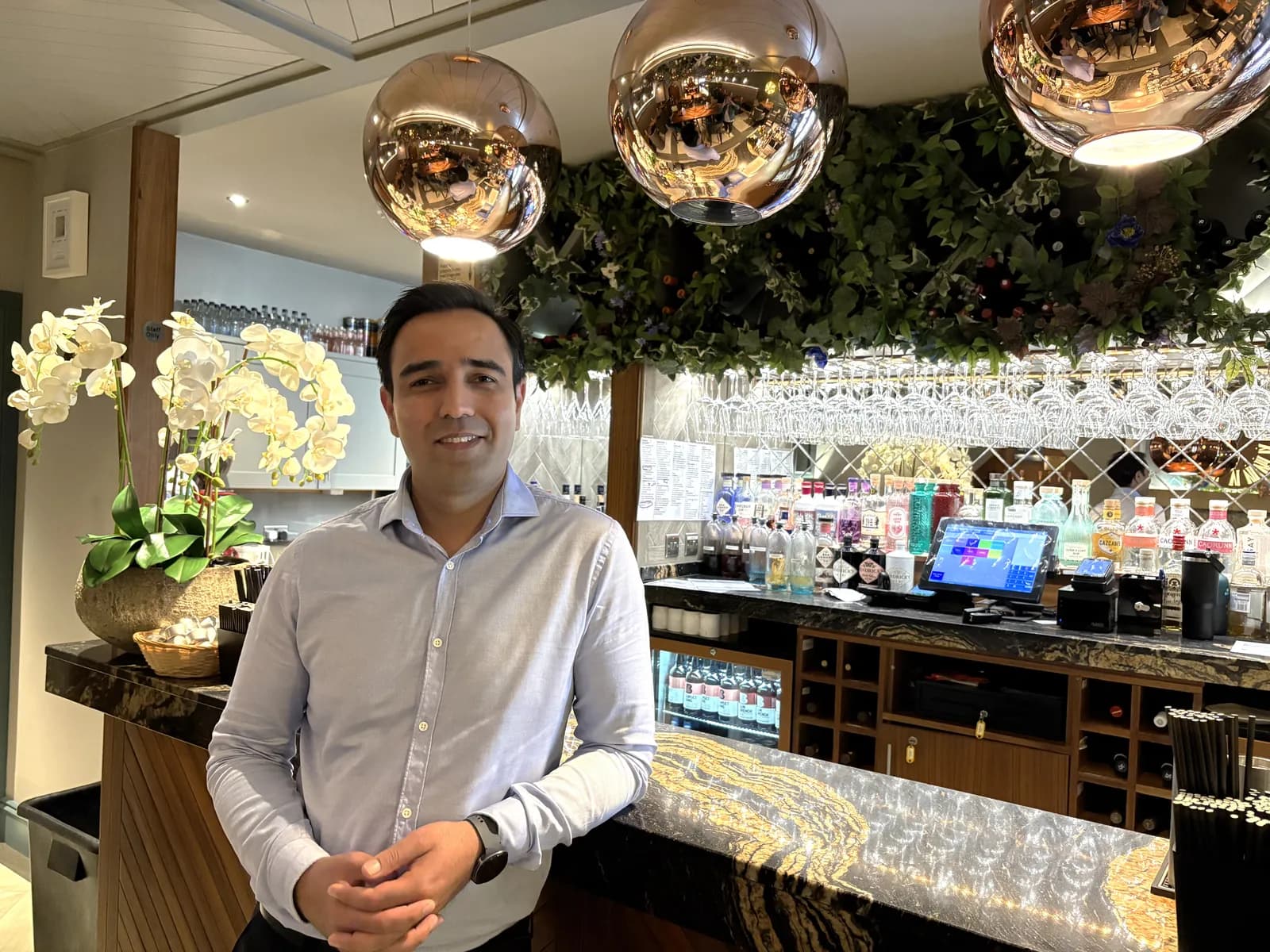 A man in a light blue button-down shirt smiles while standing in front of a bar with a large orchid plant and two hanging copper light fixtures.