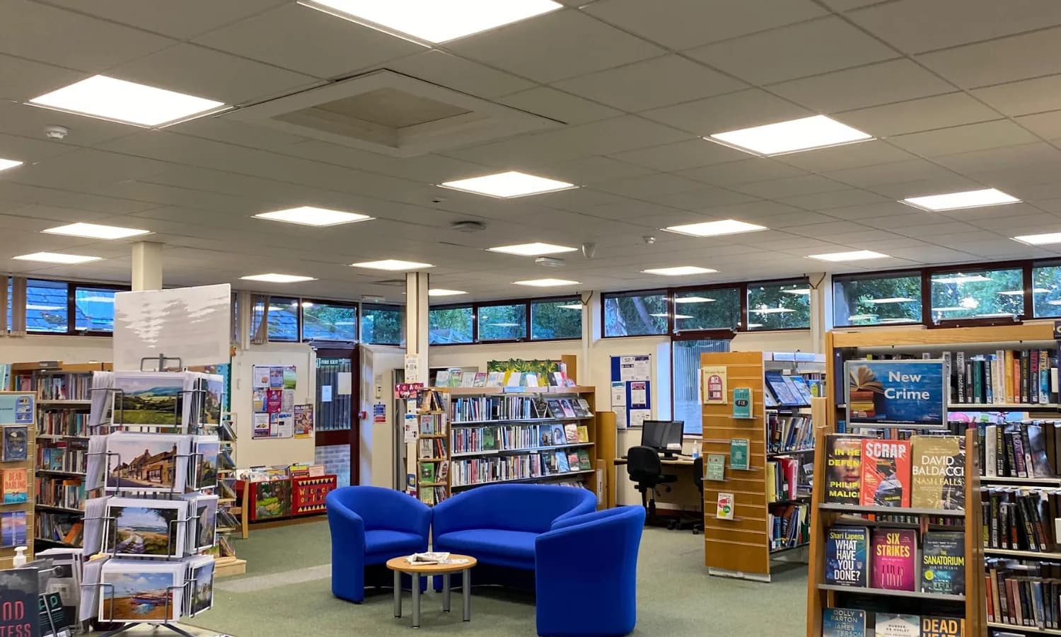 A brightly lit library interior with blue armchairs and a sofa around a small table, surrounded by bookshelves and displays.