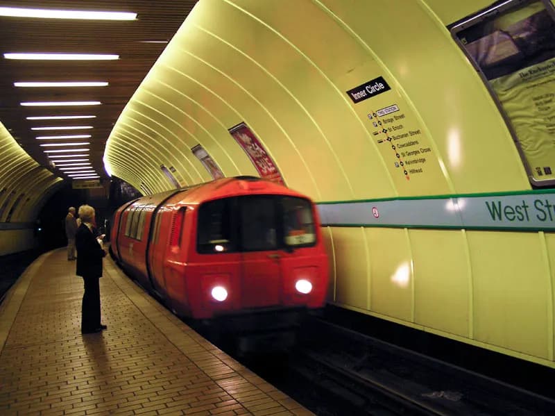 A red subway train with bright headlights approaches a station platform where a person waits.