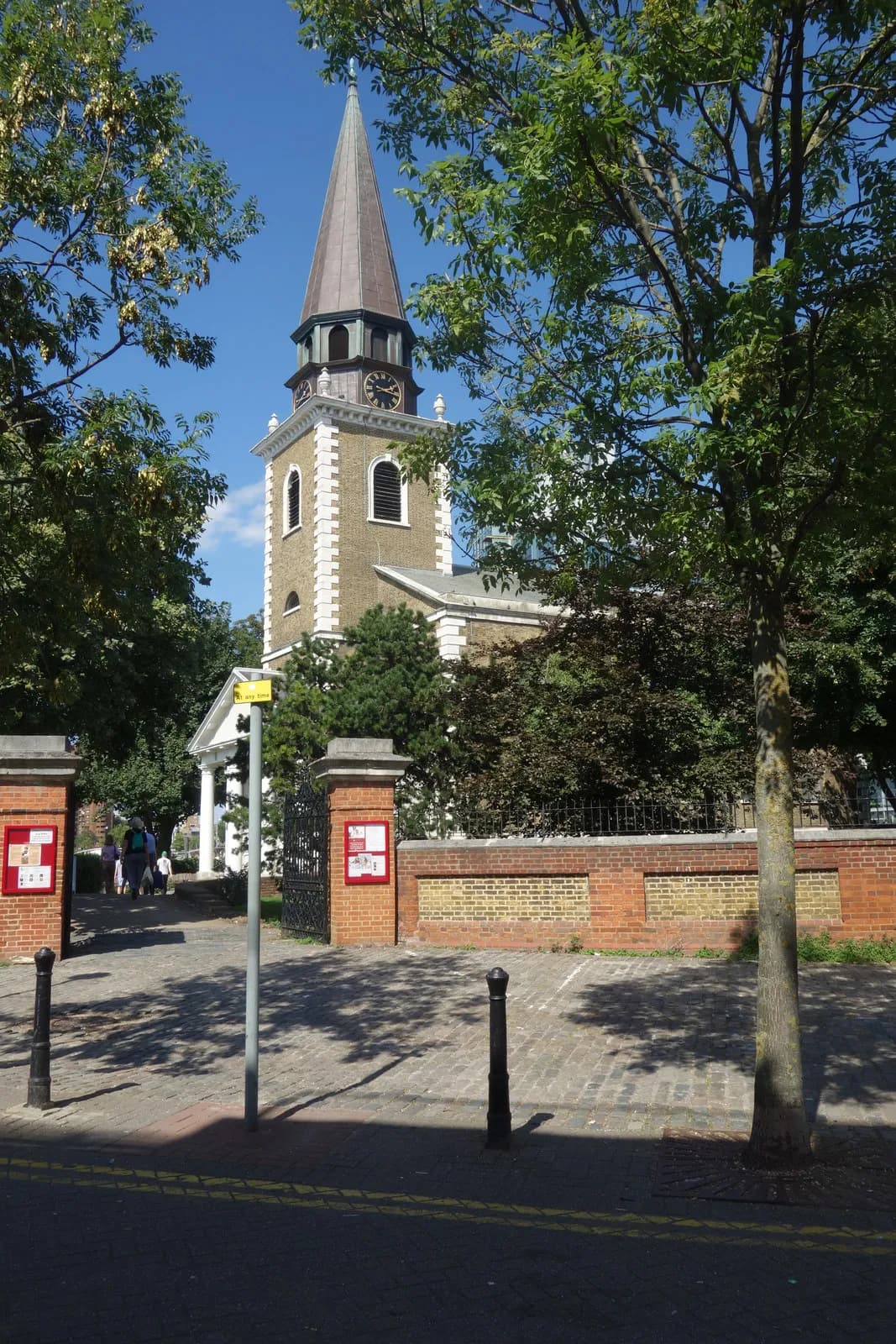 A tall church with a prominent clock tower and spired roof stands amidst green trees and a brick wall with an open gate, all under a clear blue sky.
