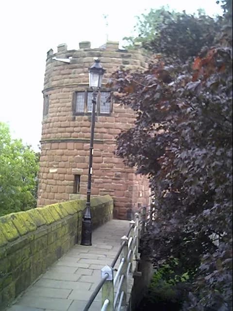 A stone bridge connecting to a round, stone tower with small windows, partially obscured by dark foliage on the right.