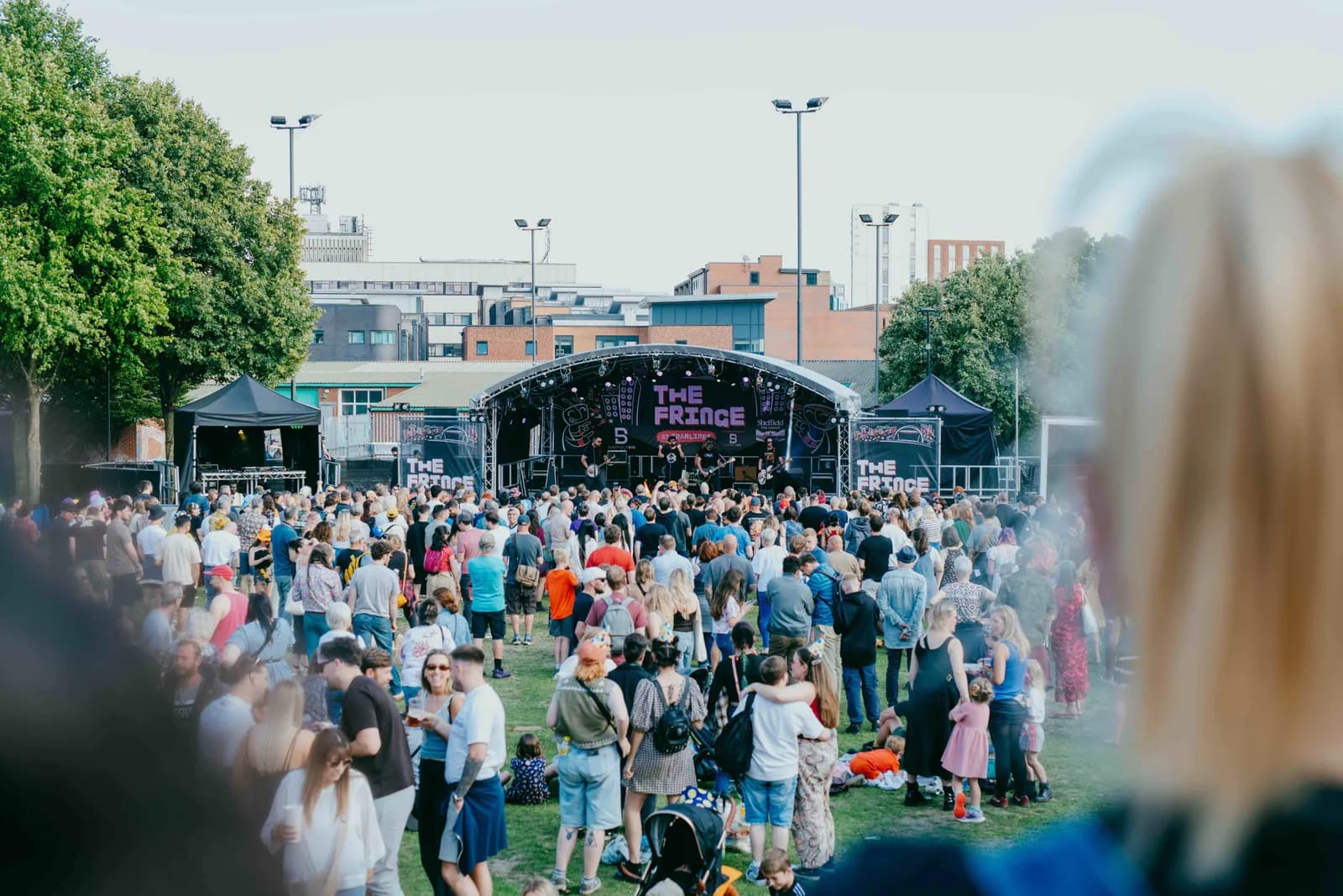 A large outdoor music festival crowd enjoys a band performing live on a brightly lit stage under a clear sky.