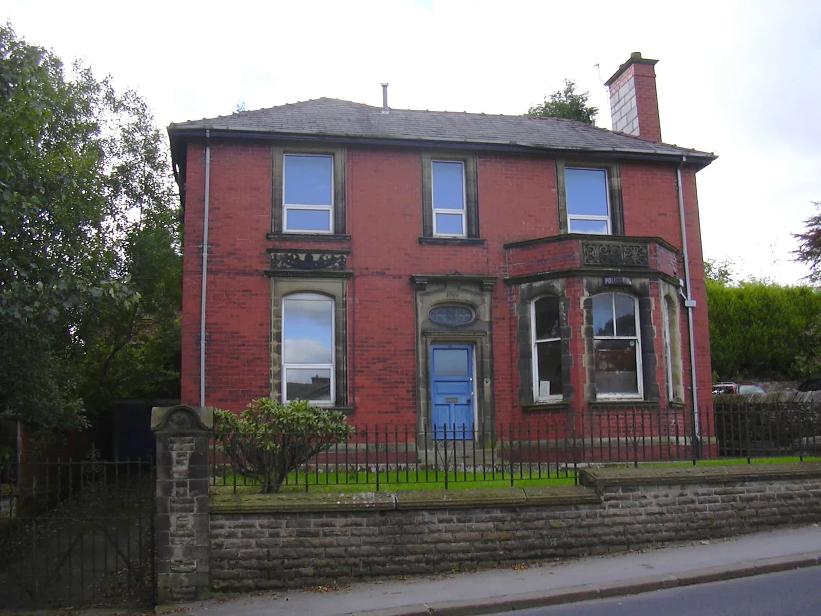 A large, two-story house with red brick walls, multiple windows, a prominent bay window, and a blue front door, surrounded by a stone wall and metal fence with green lawn.
