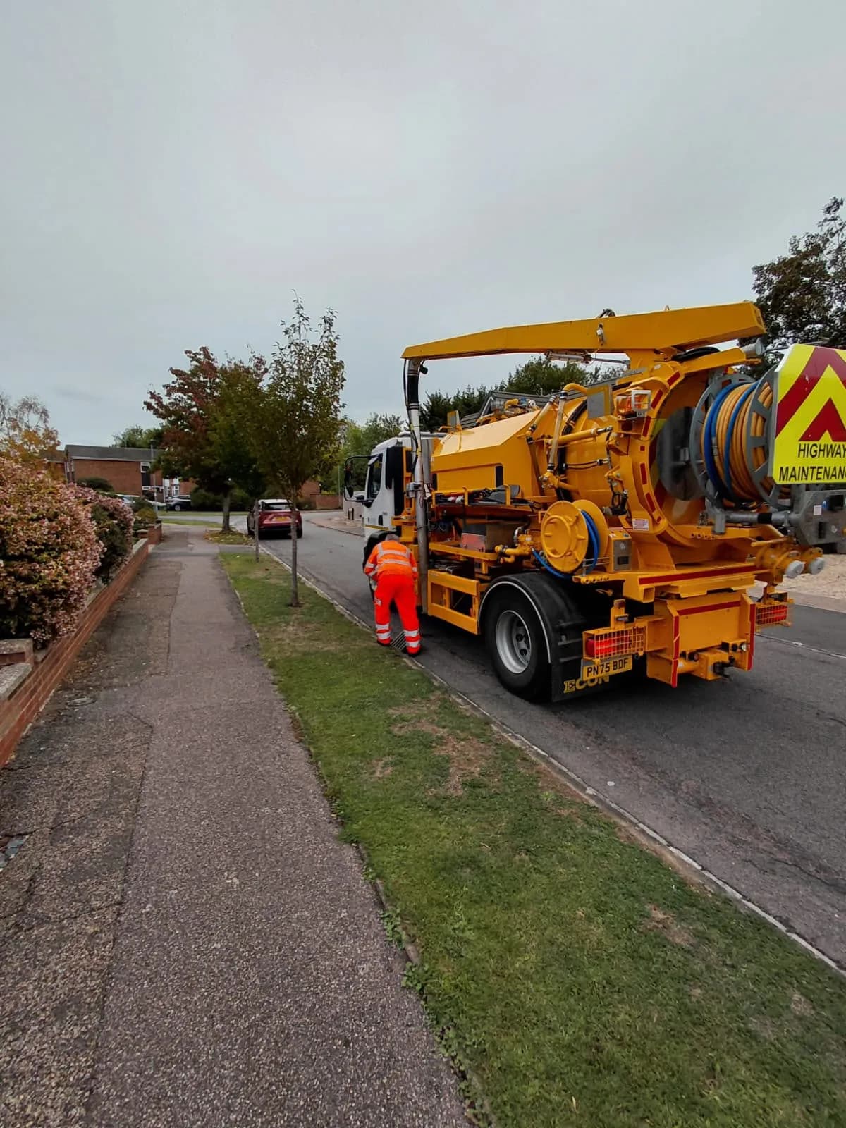 A highway maintenance worker in an orange uniform attending to a bright yellow utility truck with a large hose reel on the back, parked on the side of a residential street.