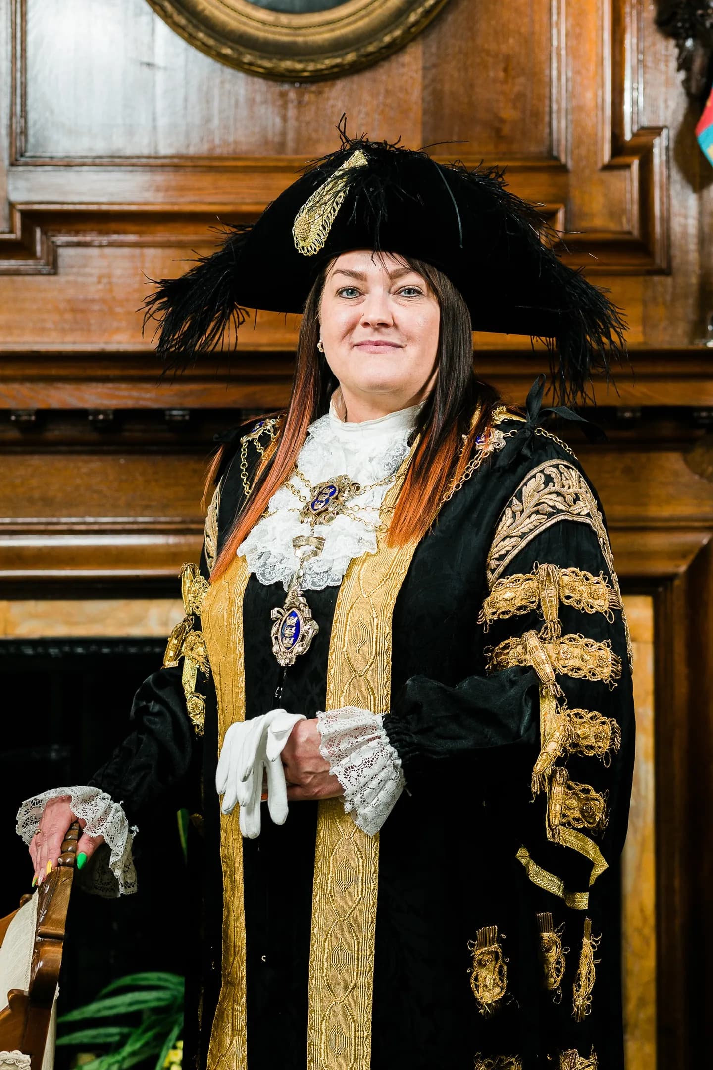 A woman in elaborate ceremonial robes with a feathered hat stands in front of a wooden wall.