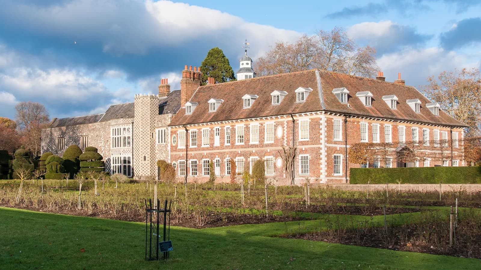 A grand, red brick manor house with white trim and multiple windows sits under a partly cloudy sky, with green lawns and garden beds in the foreground.