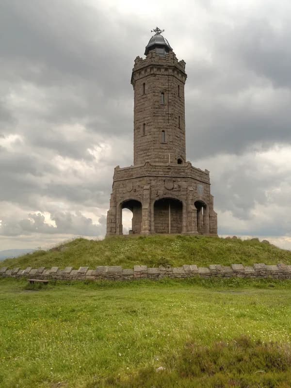 A tall stone tower with arched openings at its base stands on a grassy hill under a cloudy sky.
