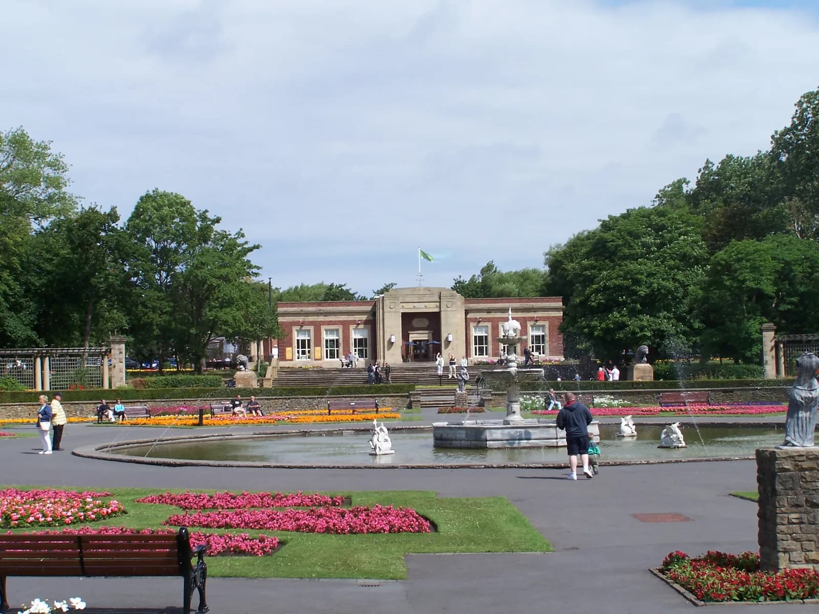 A public park featuring a large building with stairs, a pond with fountains, colorful flower beds, trees, and several people walking and sitting.