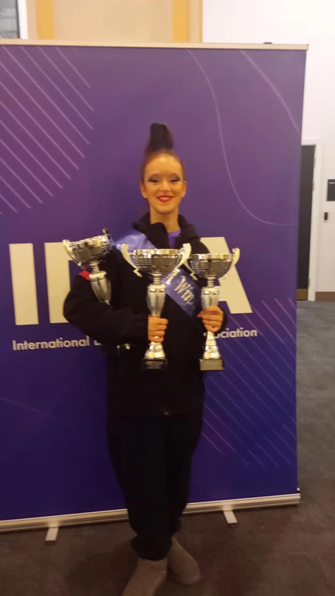 A young person with bright makeup and an elaborate hairstyle stands proudly, holding three trophies, in front of a purple banner.