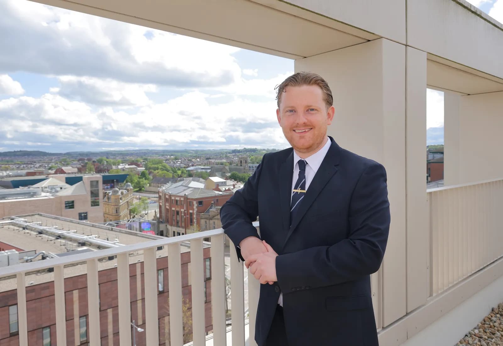 A man in a suit and tie stands on a rooftop balcony with a cityscape and cloudy sky in the background.