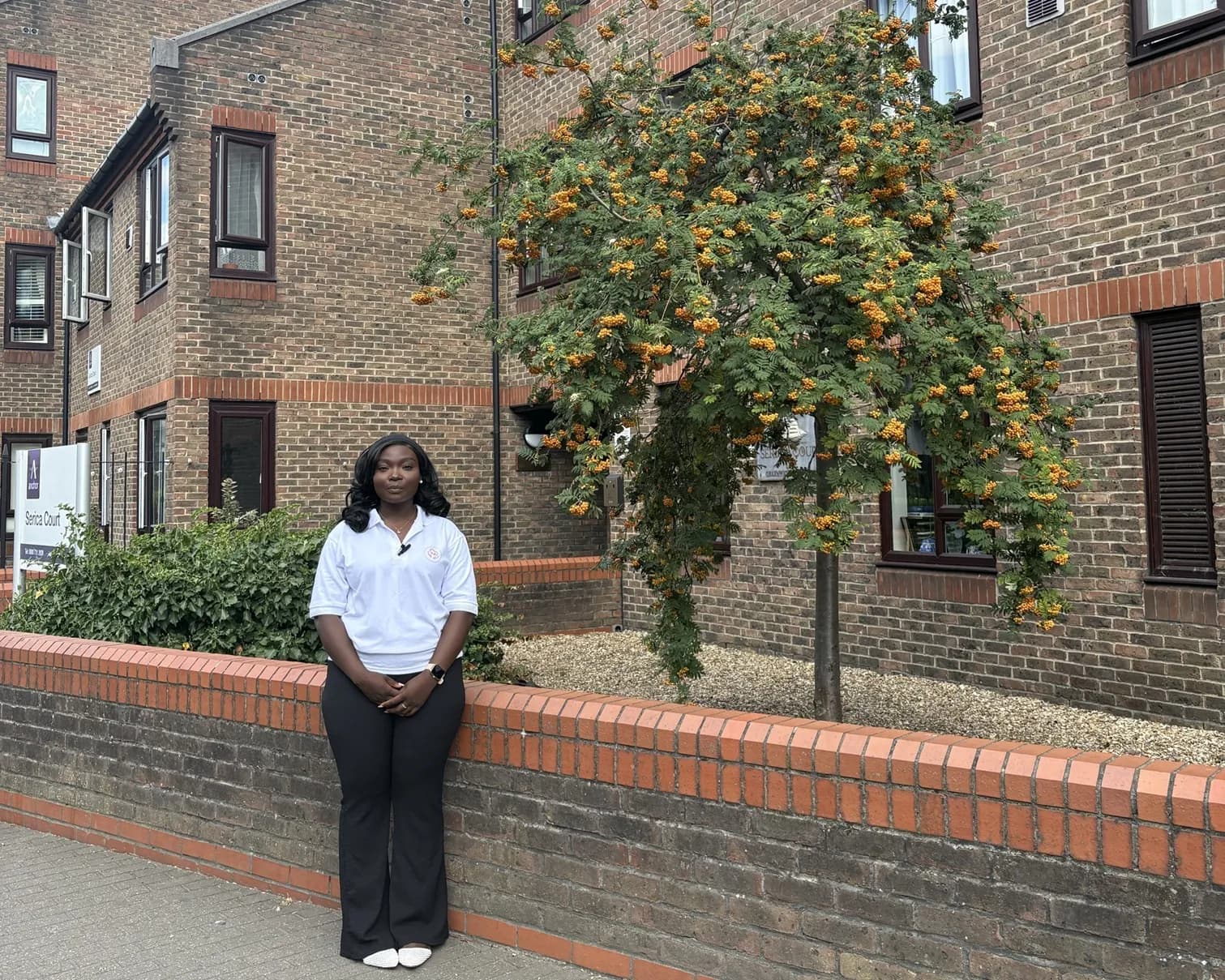 A woman stands in front of a brick building with a tree next to her.