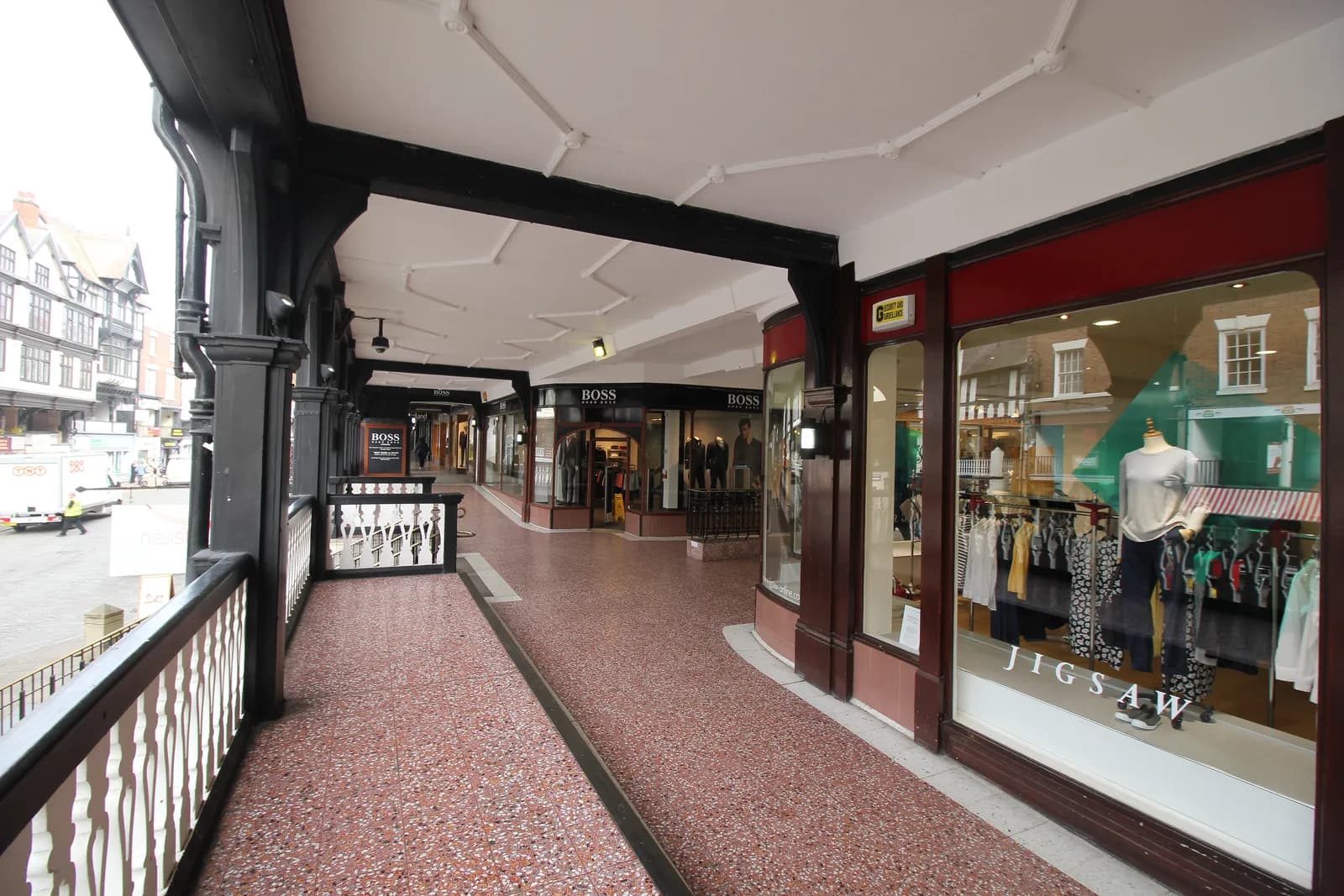 A long, covered walkway with a dark wooden railing on the left and various storefronts, including a Jigsaw clothing store, on the right.