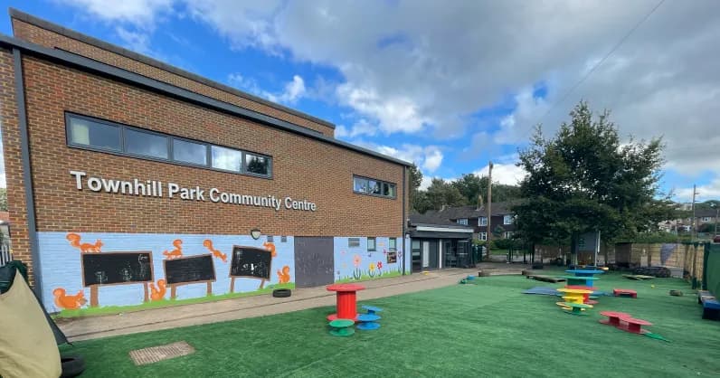 A Townhill Park Community Centre building with brick walls, grey windows, and a mural of squirrels and chalkboards. A bright green artificial turf playground with colorful equipment is in front of the building under a blue sky with white clouds.