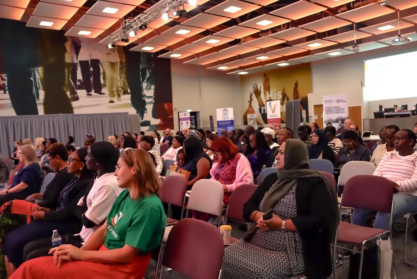 A diverse group of people, including men and women, are seated and looking forward in a conference room, with some wearing headscarves and others in casual attire, against a backdrop of large wall art.
