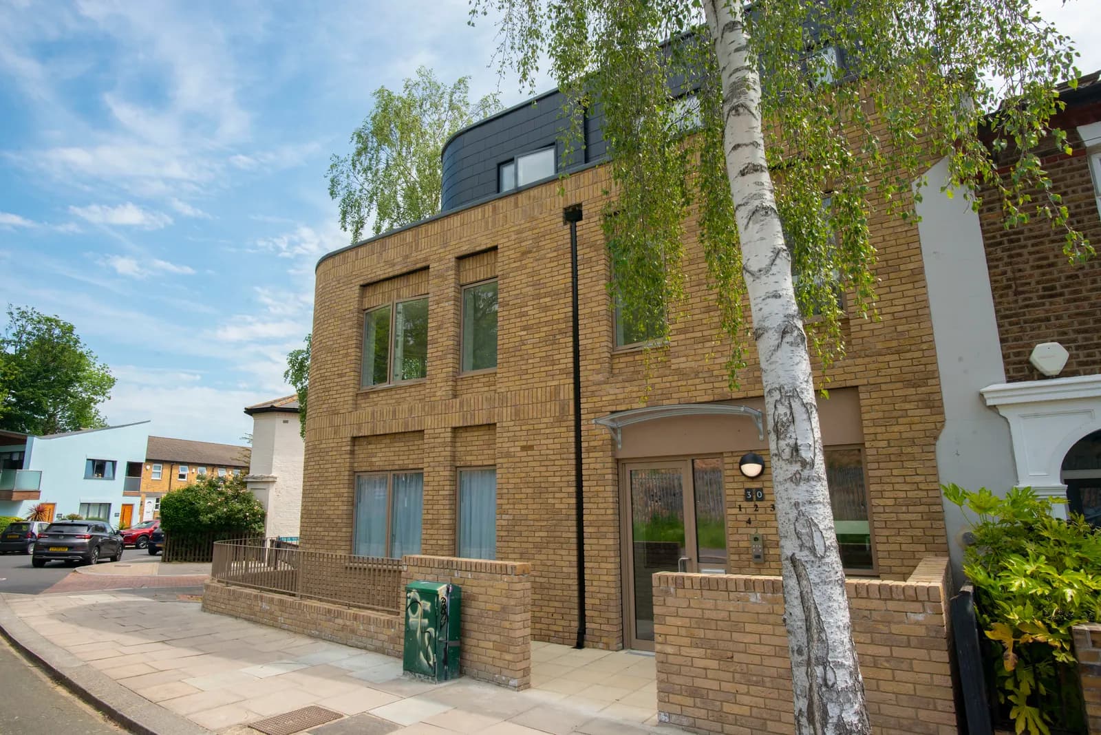 A modern brick building with a dark grey curved roof extension stands next to a birch tree on a sunny day.