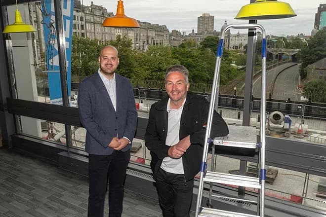 Two men standing inside a modern building with large windows overlooking a city street and industrial construction.