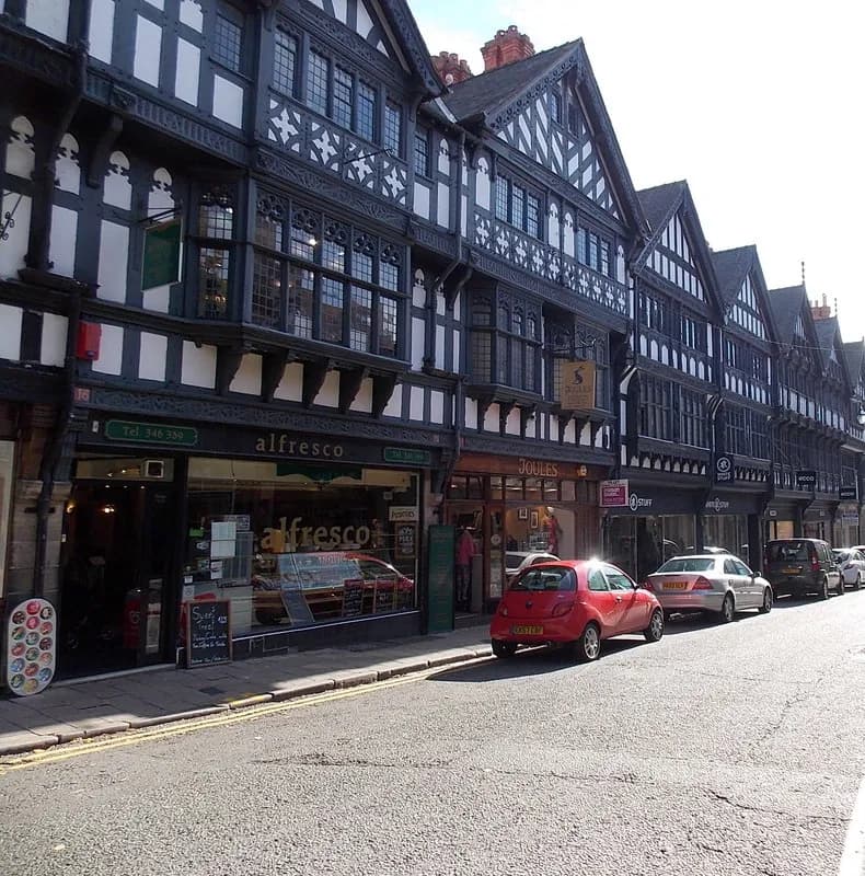 A line of historic black and white timber-framed buildings with shops on the ground floor, and several cars parked along the street on a sunny day.
