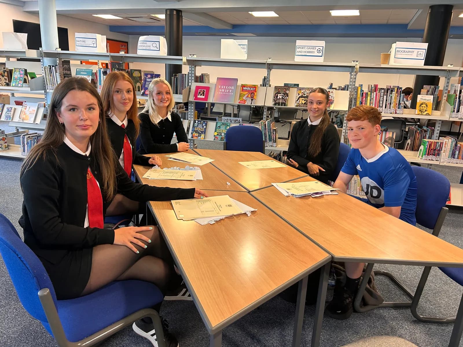 Six high school students, five girls and one boy, are sitting around a table in a library with papers spread out before them.