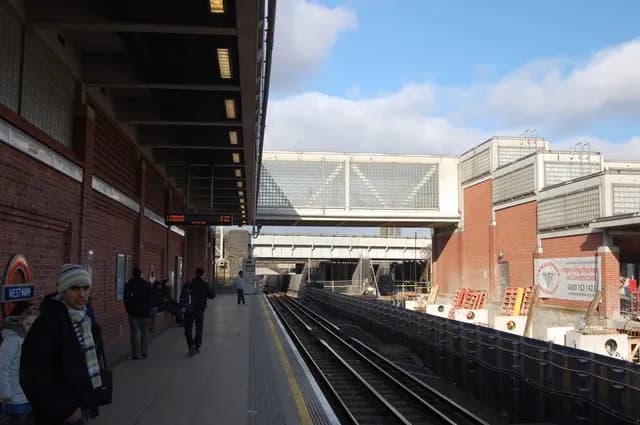 A man wearing a striped beanie and scarf stands on a subway platform with train tracks and a pedestrian bridge overhead.