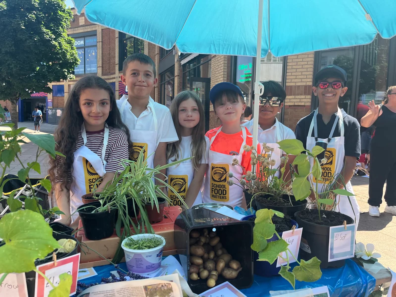 Six children, three boys and three girls, stand behind a table filled with potted plants and produce under a blue umbrella at an outdoor market.