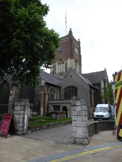A historic stone church with a tall clock tower, visible through the branches of a large green tree, with a white van parked near the building on the right.