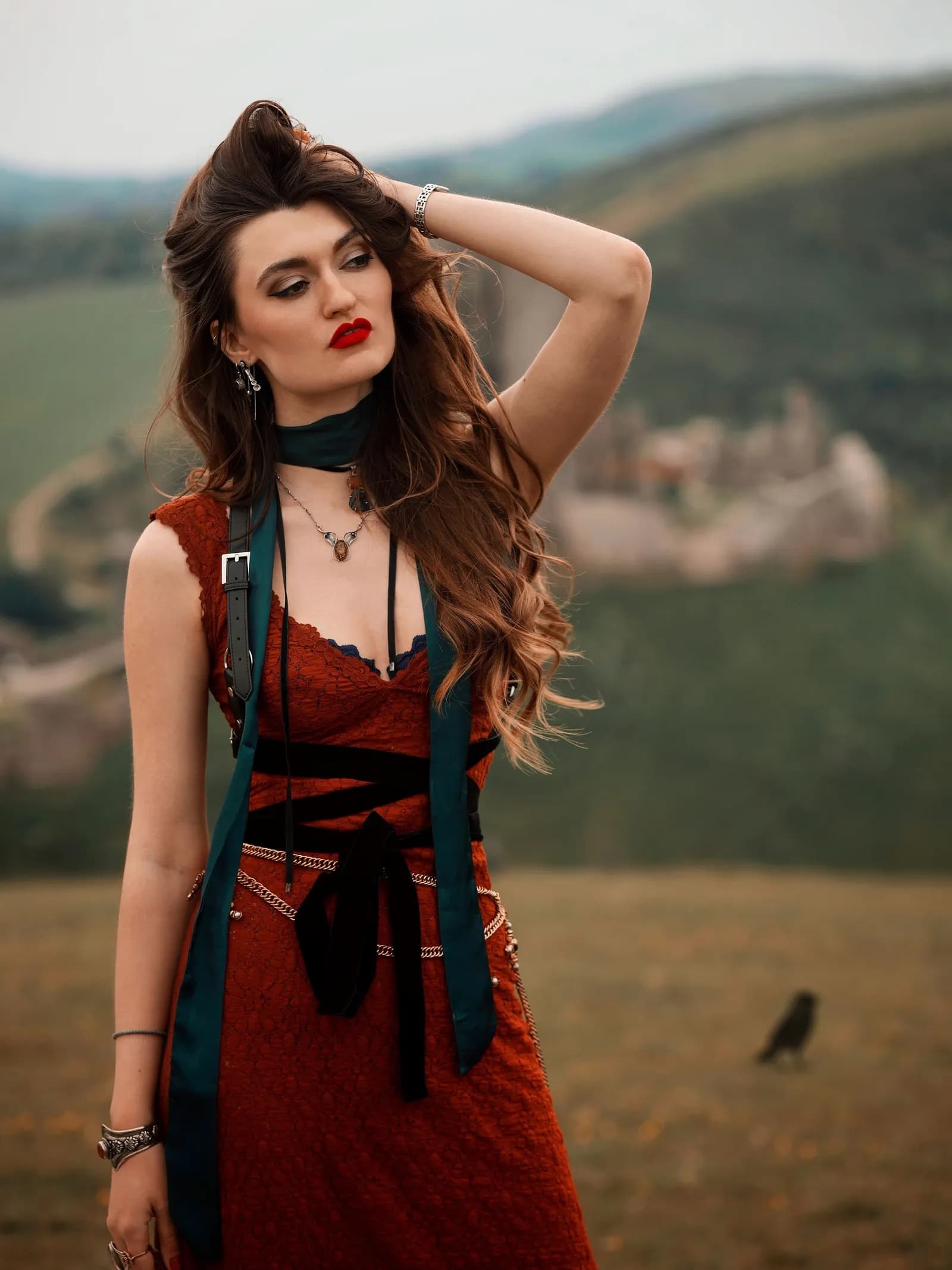 A woman with long wavy brown hair, wearing a red dress, green scarf, and ornate jewelry, poses with her hand in her hair against a background of rolling hills and a distant ruined castle.