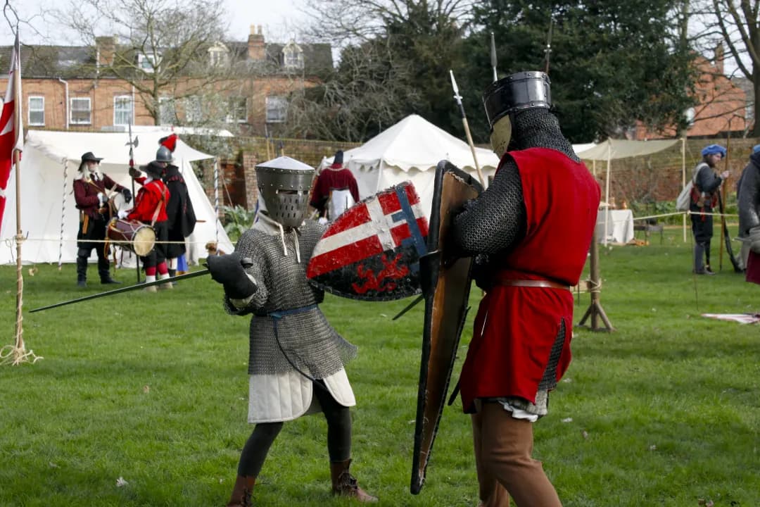 Two medieval knights in armor and shields engage in a mock battle on a grassy field, with tents and other costumed individuals in the background.