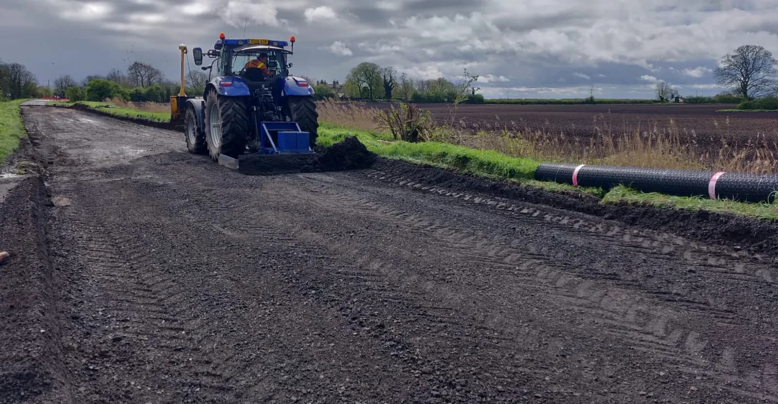 A blue tractor is seen from behind and slightly to the left, leveling a dark gravel pathway, with a field in the background under a cloudy sky.