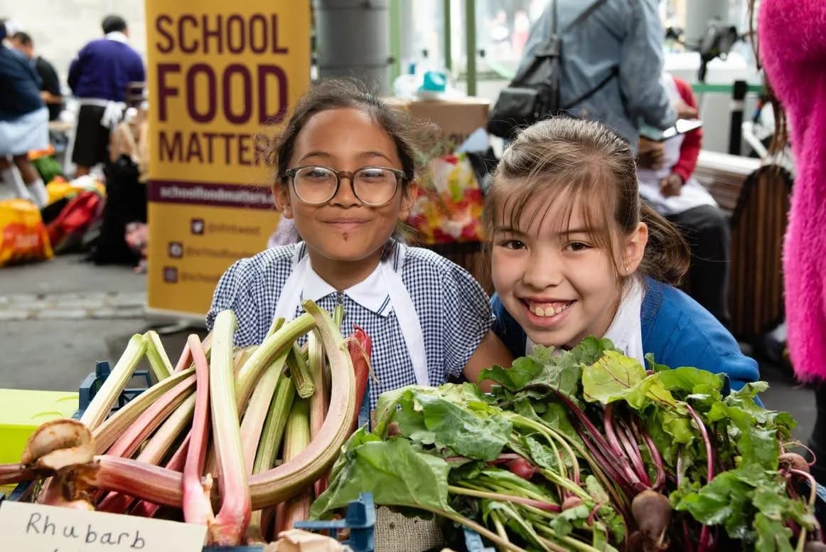 Two young girls in school uniforms smiling with fresh rhubarb and beets in front of them at an outdoor market stall with a yellow banner in the background that reads "SCHOOL FOOD MATTERS."