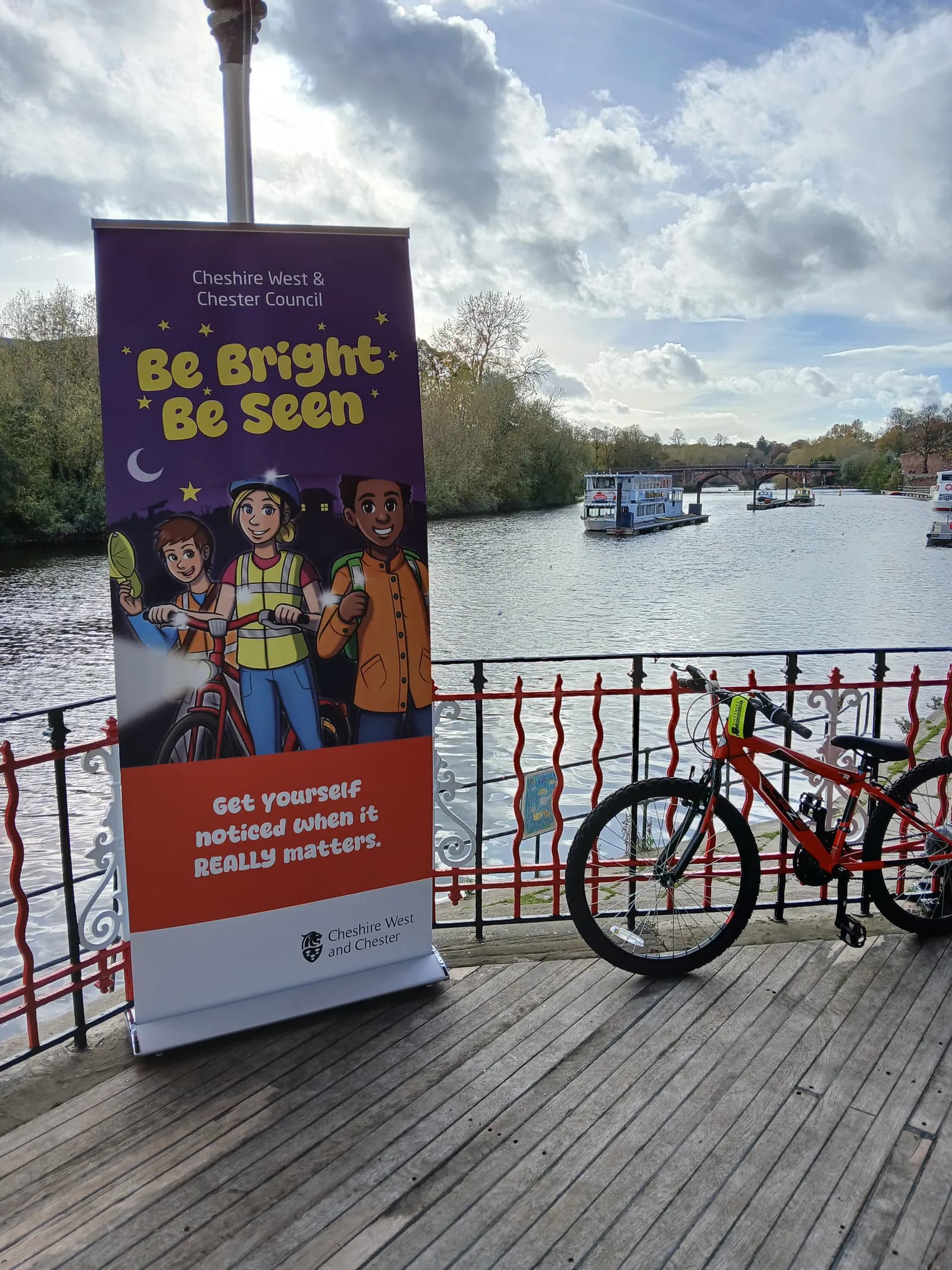 A banner with the slogan "Be Bright Be Seen" featuring three cartoon children wearing reflective gear stands beside a bicycle on a wooden deck overlooking a river with boats.