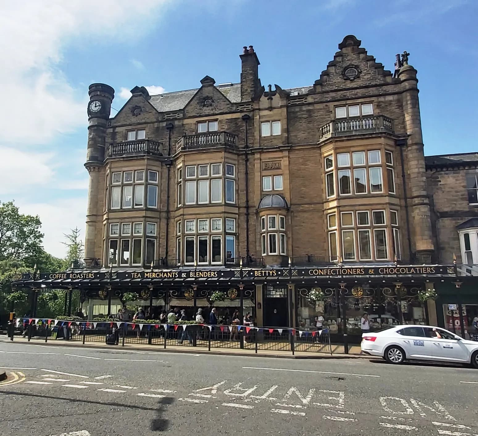A grand, multi-story stone building with ornate bay windows, balconies, and a clock tower, housing a cafe with outdoor seating and a white car driving past on the street.