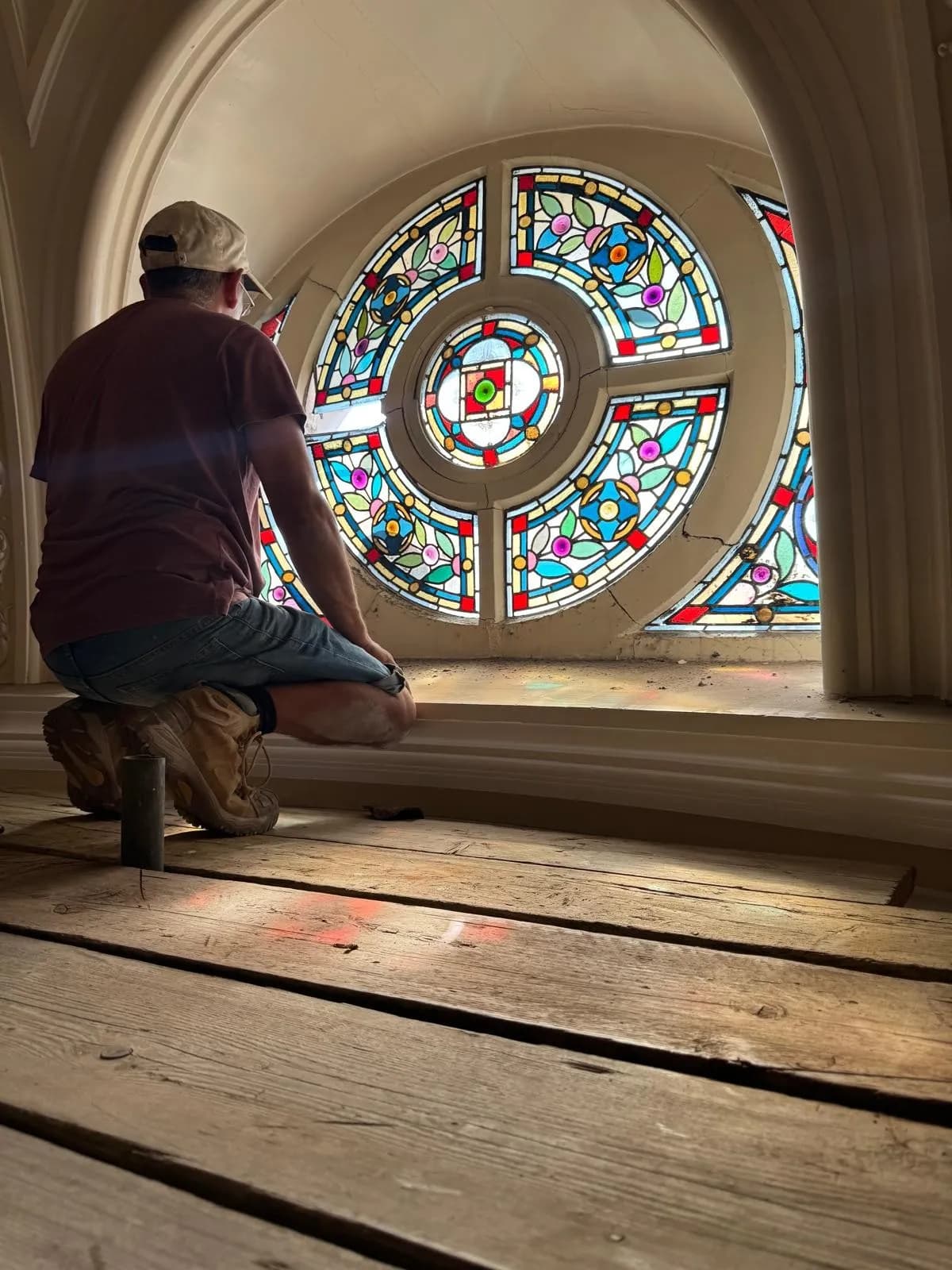 A person kneeling on a wooden floor, looking out of a large, circular stained glass window with vibrant colors and intricate patterns.